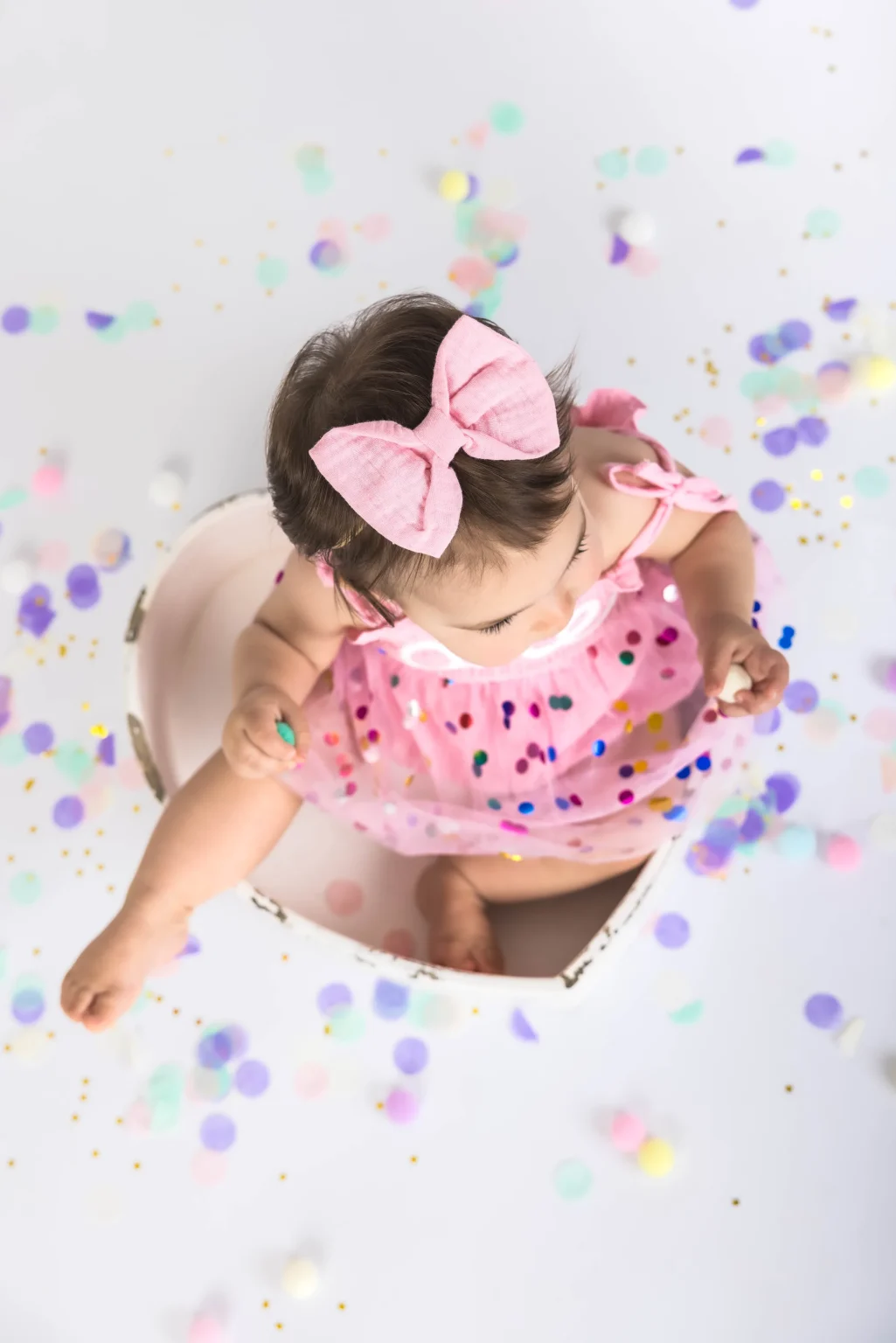 A baby wearing a pink dress with a matching pink bow sits in a heart-shaped container surrounded by colorful confetti.