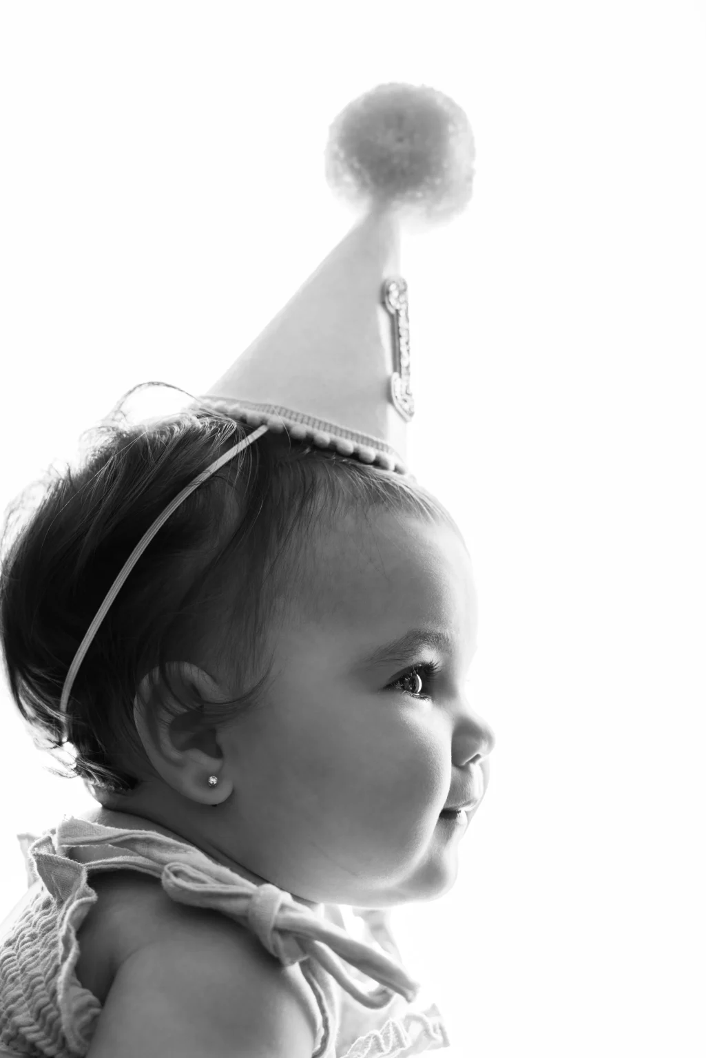 Black and white side profile of a baby wearing a party hat, looking forward and smiling.