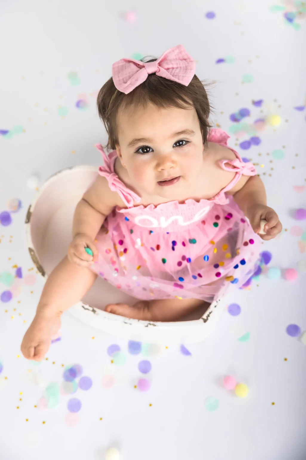 A baby wearing a pink dress and bow sits in a heart-shaped bowl surrounded by colorful confetti and pom-poms on a white background.