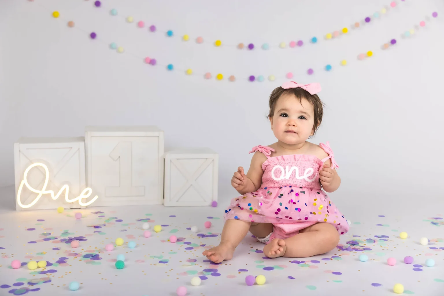 A baby wearing a pink dress with "One" on it sits among colorful confetti and pom-poms, with decorated boxes and a light-up "One" sign in the background.