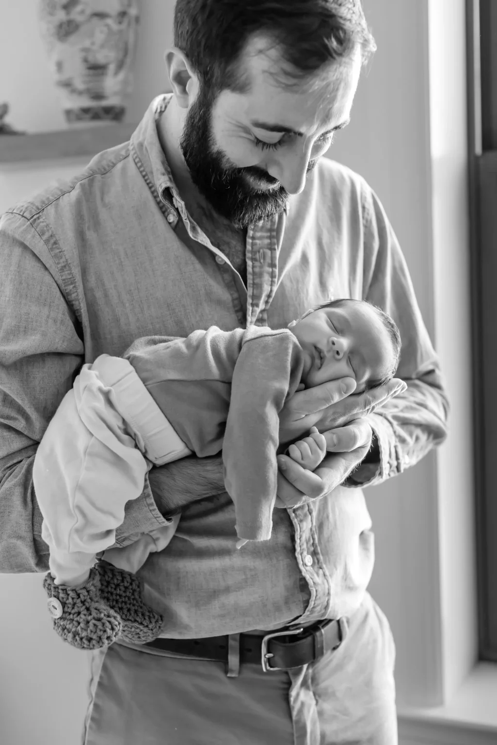 A man holding a sleeping newborn baby in his arms, both dressed in casual clothes, indoors by a window.