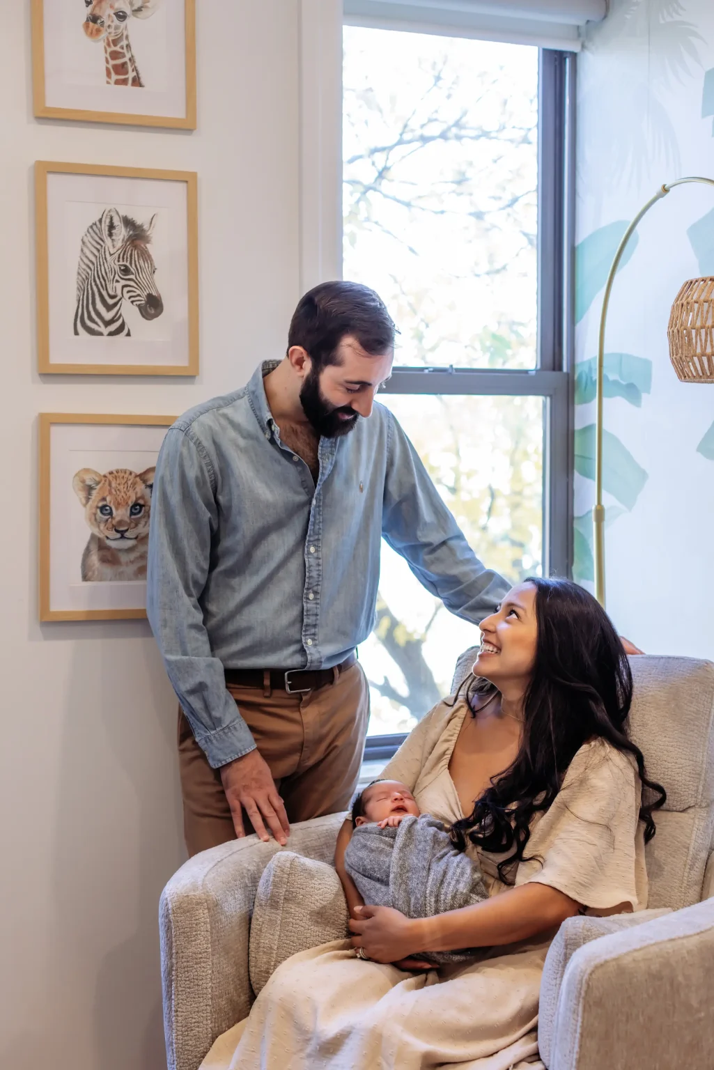 A man stands beside a woman sitting in a chair holding a baby. They are in a bright room with animal artwork and a large window in the background.