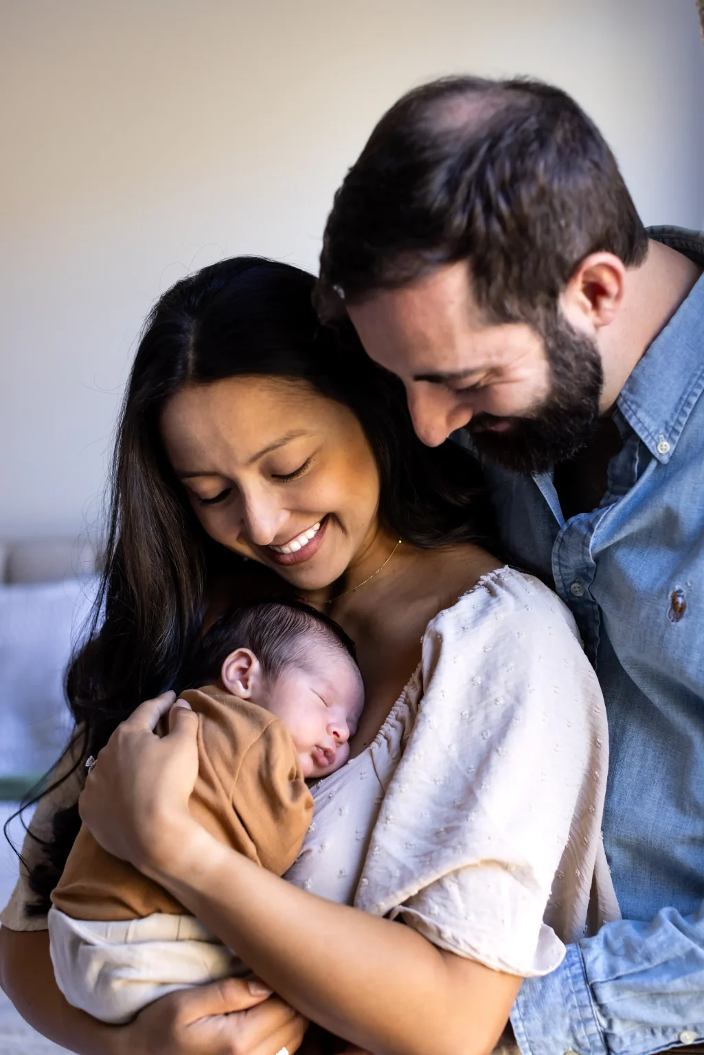 A woman holds a sleeping baby in her arms while a man stands close beside them, both adults smiling and looking down at the baby.