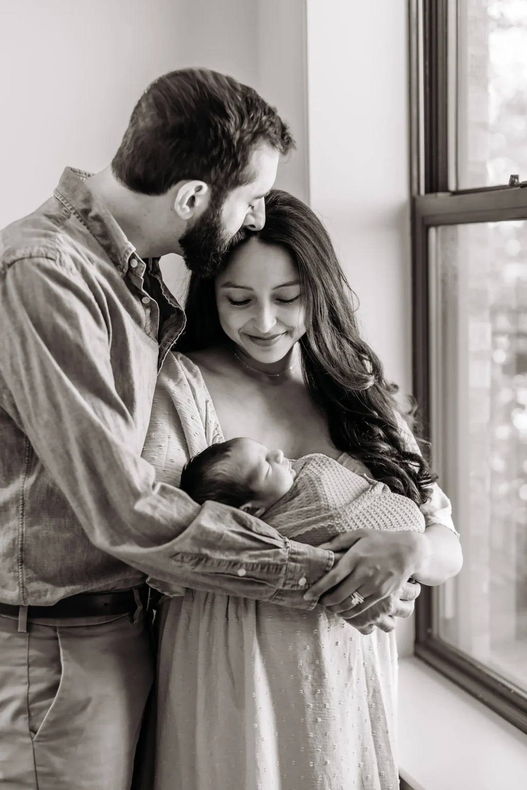 A man and woman stand by a window, smiling while holding a sleeping baby wrapped in a blanket. The man gently kisses the woman's head.