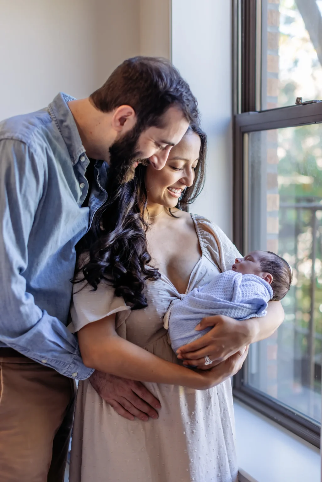 A couple stands by a window, smiling down at a newborn wrapped in a blanket held in the woman’s arms.