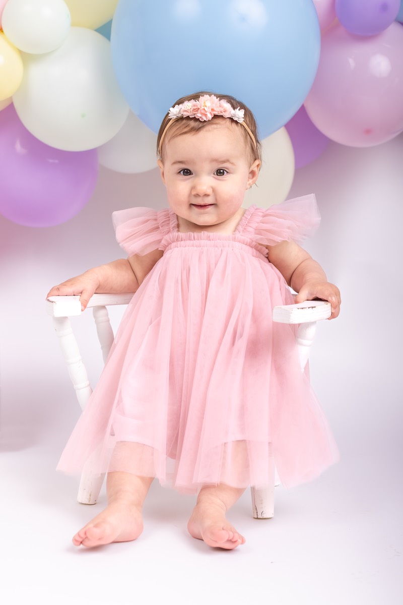 A baby wearing a pink dress and flower headband sits on a white chair with colorful balloons in the background.