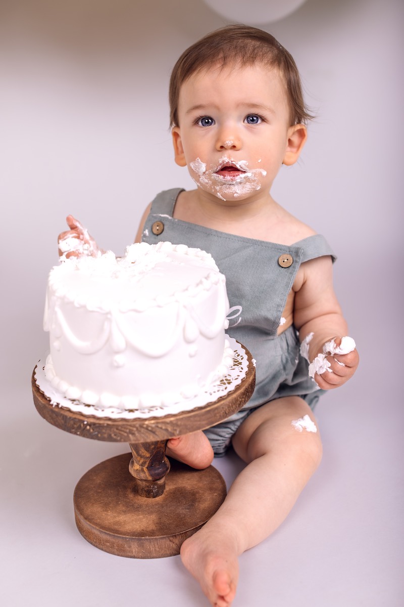 A baby in a grey romper sits on the floor with a white frosted cake, touching and eating the cake, with frosting on their face and hands.