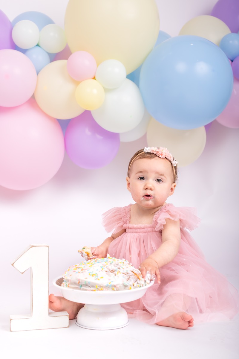 A baby in a pink dress sits next to a cake and a large number one, with colorful balloons in the background.
