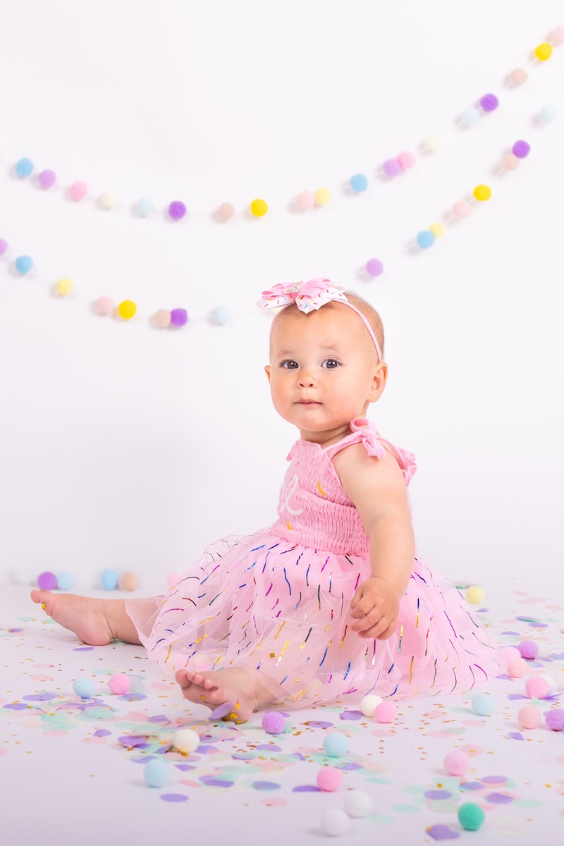 A baby wearing a pink dress and headband sits on the floor surrounded by colorful pom-poms, with pastel garlands hanging in the background.