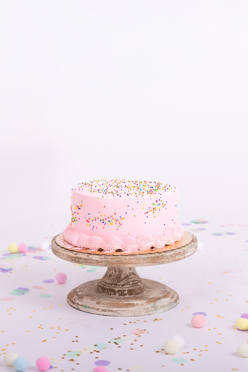 A pink frosted cake with colorful sprinkles sits on a wooden cake stand, surrounded by confetti and small pastel pom-poms on a white background.