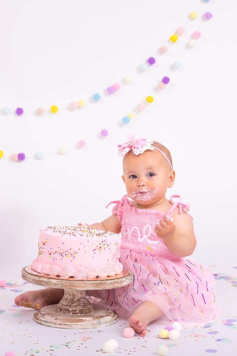 A baby in a pink dress sits on the floor with a pink frosted cake, some frosting on her face, and colorful decorations in the background.