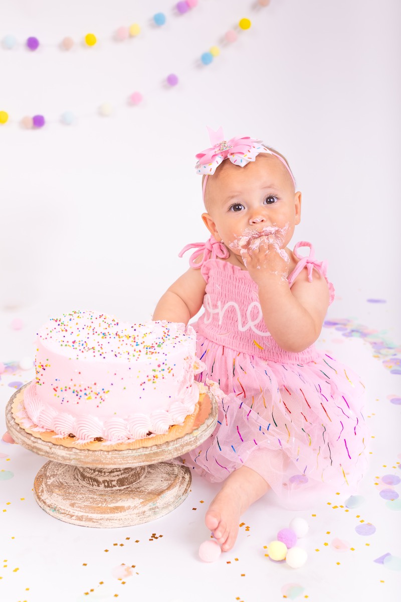 A baby in a pink dress sits next to a pink frosted cake, with frosting on her hand and mouth, surrounded by confetti and pastel decorations.