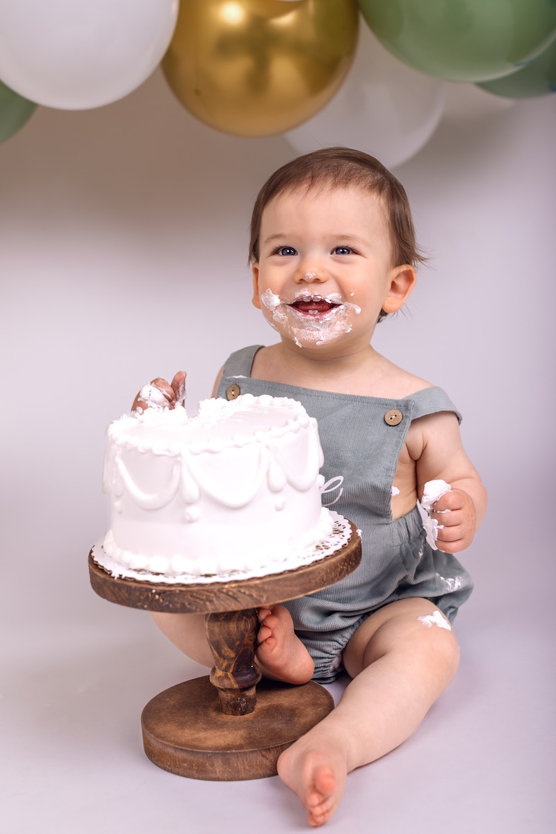 A baby in a grey romper sits on the floor with a frosted cake, smeared with cake and frosting on their face and hands. Balloons are in the background.