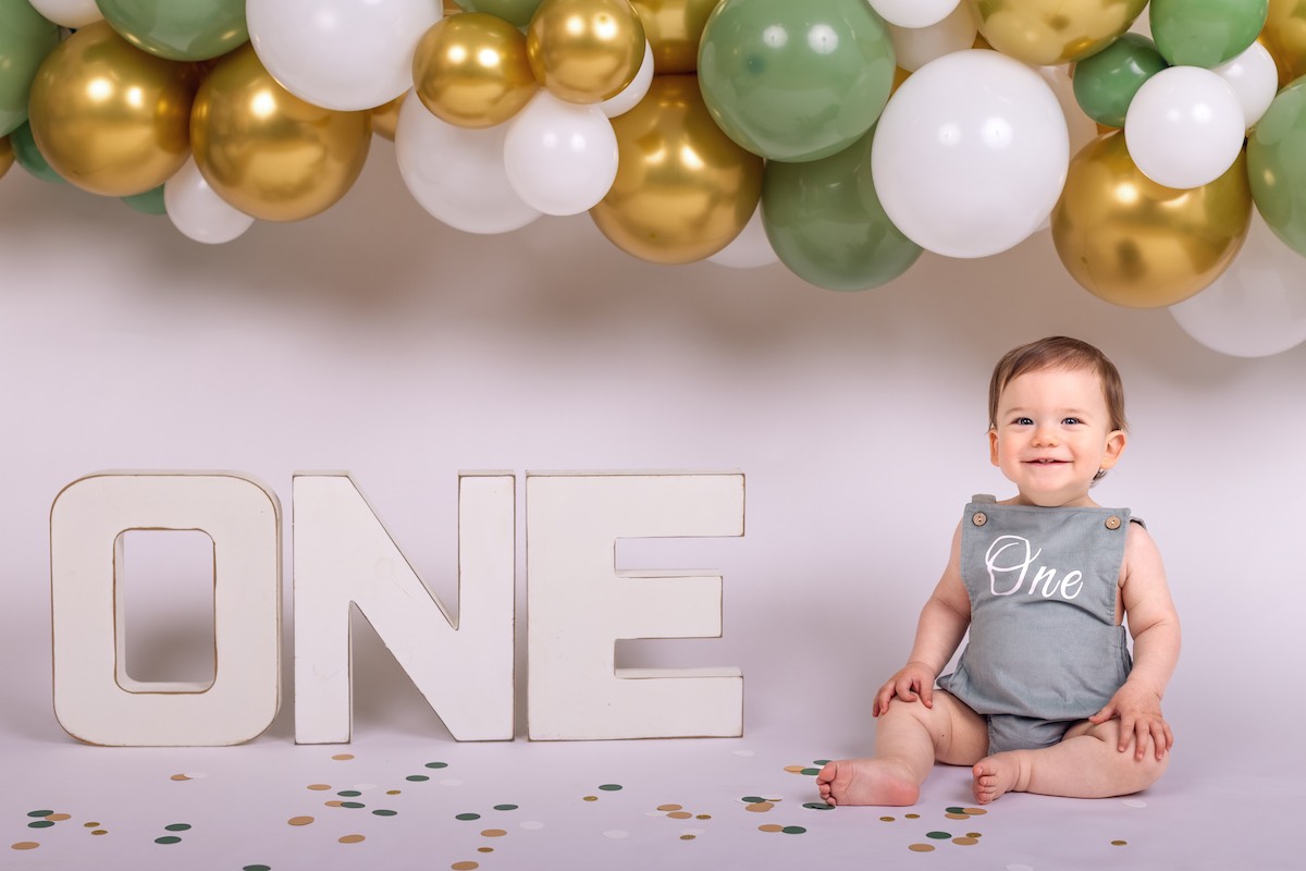 A baby wearing a grey romper with "One" printed on it sits beside large letters spelling "ONE," with green, gold, and white balloons in the background.