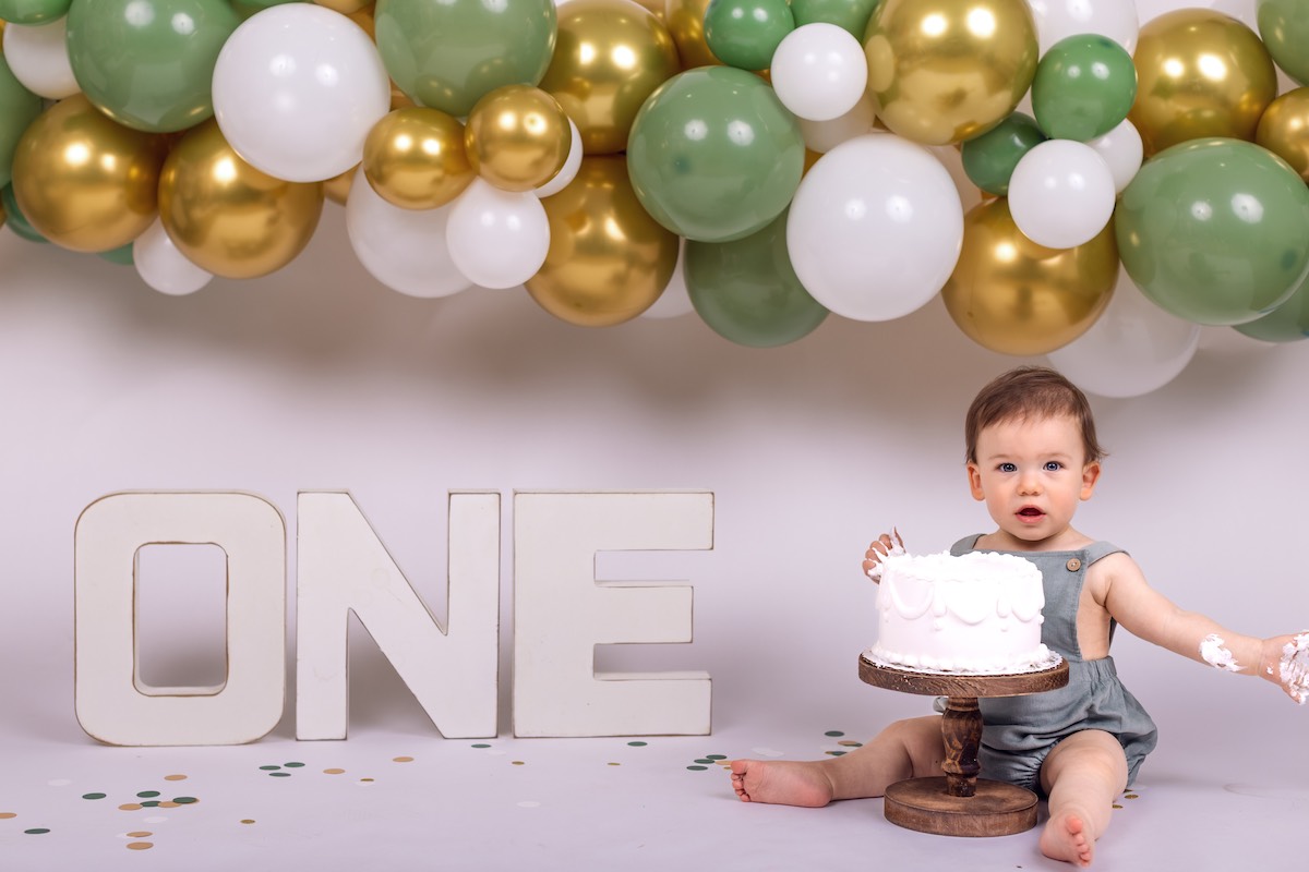 A baby sits next to a cake with "ONE" letters and gold, green, and white balloons in the background, celebrating a first birthday.