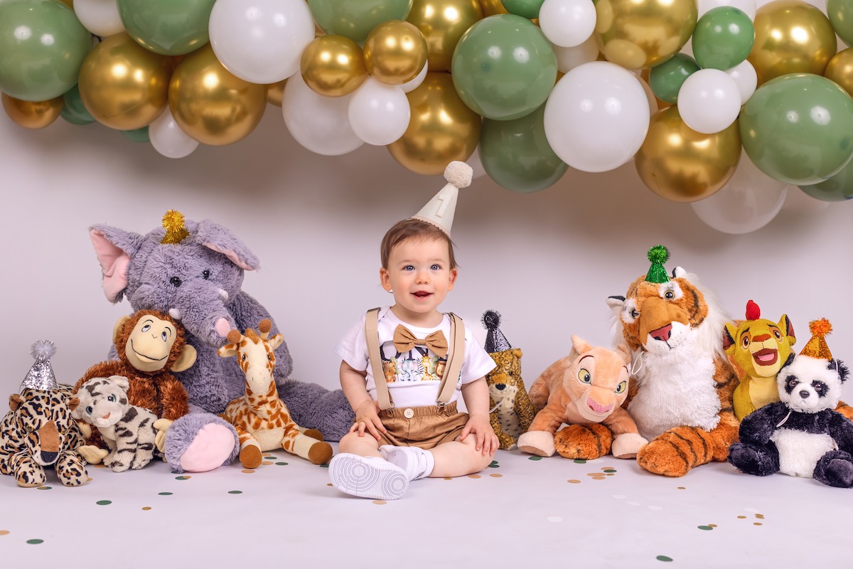 A young child wearing a party hat sits on the floor surrounded by stuffed animals and festive balloon decorations in green, gold, and white.