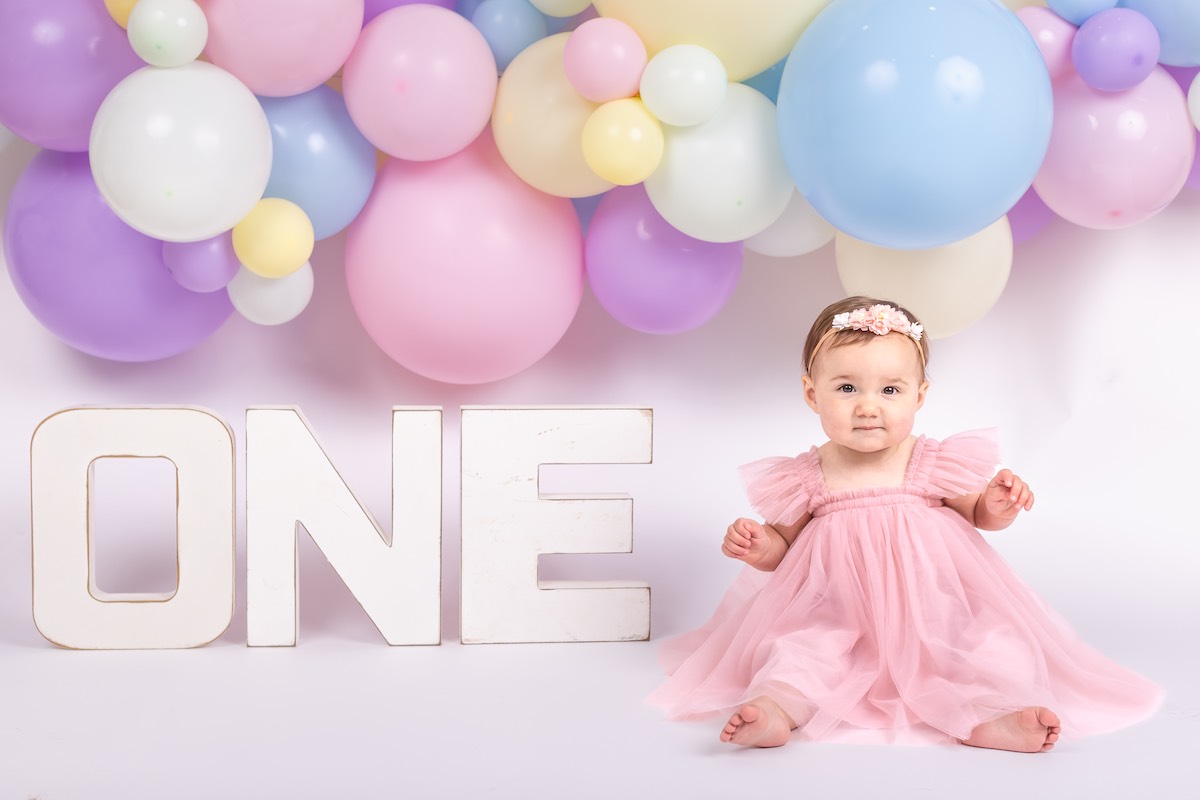 A baby in a pink dress sits next to large letters spelling "ONE" with pastel balloons in the background.