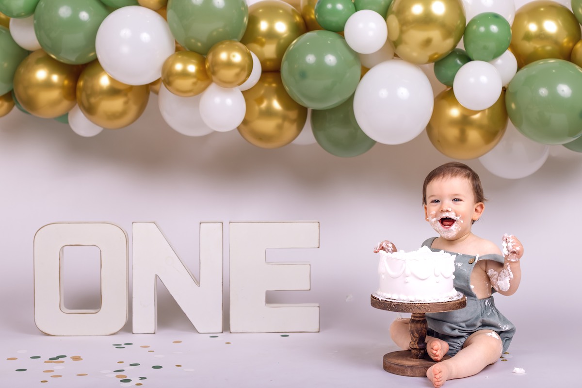 A baby with cake on their face sits by a white cake and large letters spelling "ONE," with green, white, and gold balloons in the background.