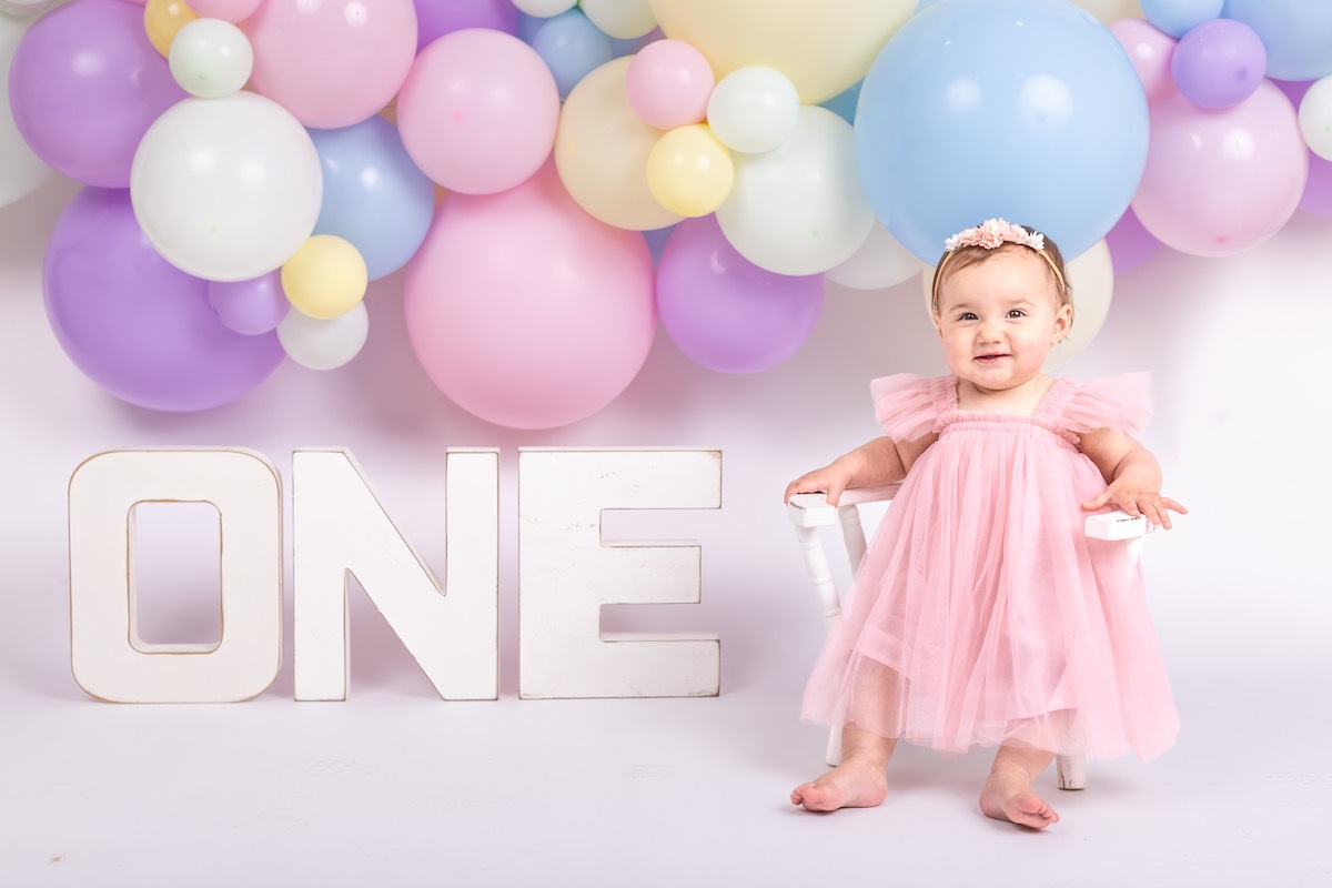 A baby in a pink dress sits on a white chair next to large white letters spelling "ONE," with pastel balloons in the background.