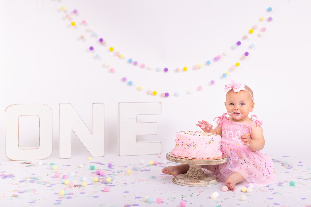 Baby in a pink dress sits next to a pink cake and large letters spelling "ONE," surrounded by pastel decorations and confetti, marking a first birthday celebration.