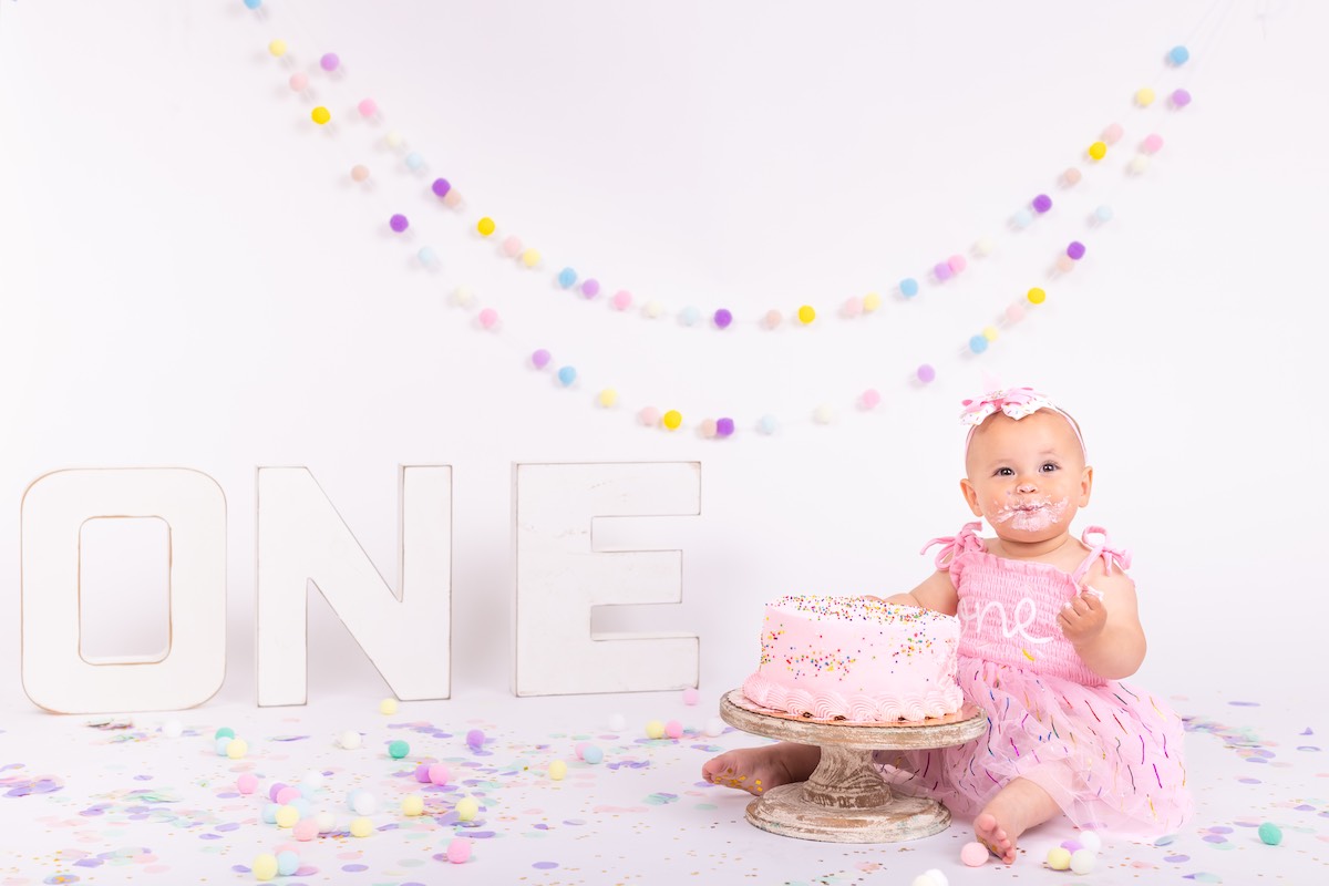 A baby in a pink dress sits beside a pink cake with “ONE” letters and pastel decorations, celebrating a first birthday.
