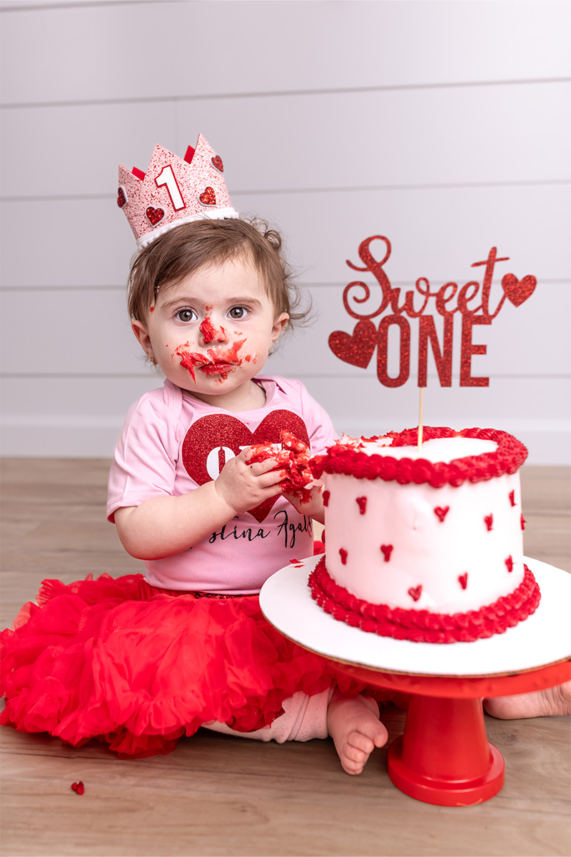 A baby in a red tutu and "1" crown sits on the floor with a frosted cake, partially smeared on her face and hands, celebrating a first birthday—captured by a Long Island photographer.