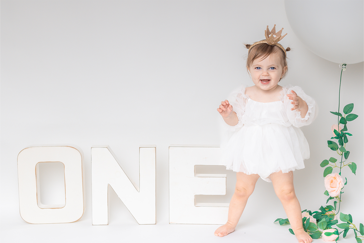 A toddler in a white outfit and gold crown stands beside large letters spelling "ONE" and a white balloon adorned with greenery and flowers, captured beautifully by a Long Island photographer.