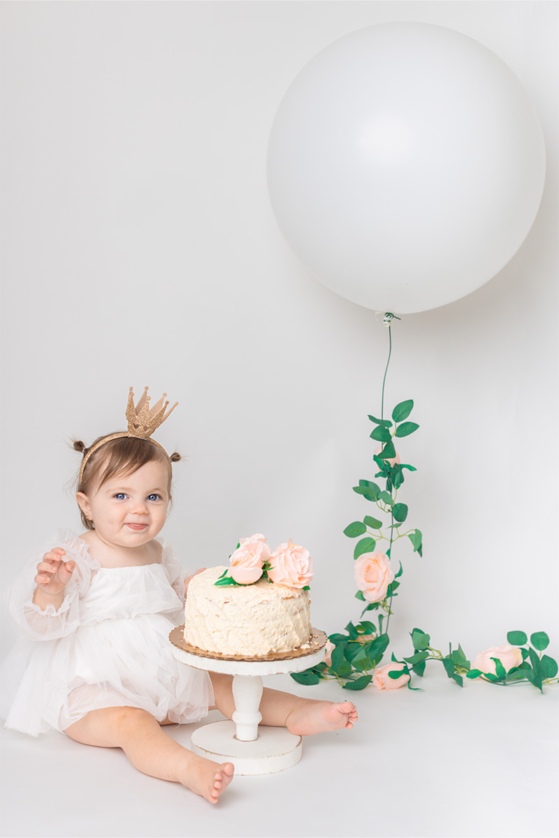 A baby wearing a crown sits barefoot next to a cake with flowers and a large white balloon decorated with greenery and roses, against a plain white background—perfectly captured by a Long Island photographer.