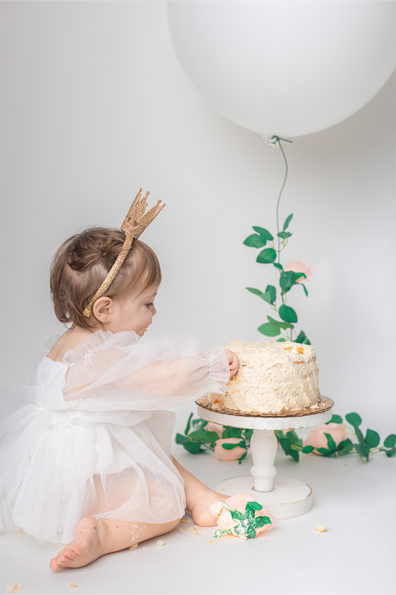 A baby in a white dress and gold crown sits on the floor, reaching into a frosted cake on a stand, with a white balloon and greenery in the background—captured beautifully by a Long Island photographer.