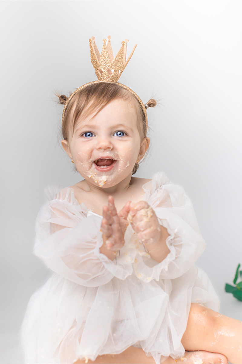 A baby wearing a white dress and a gold crown headband sits smiling with cake on her face and hands, captured by a Long Island photographer, against a plain white background.
