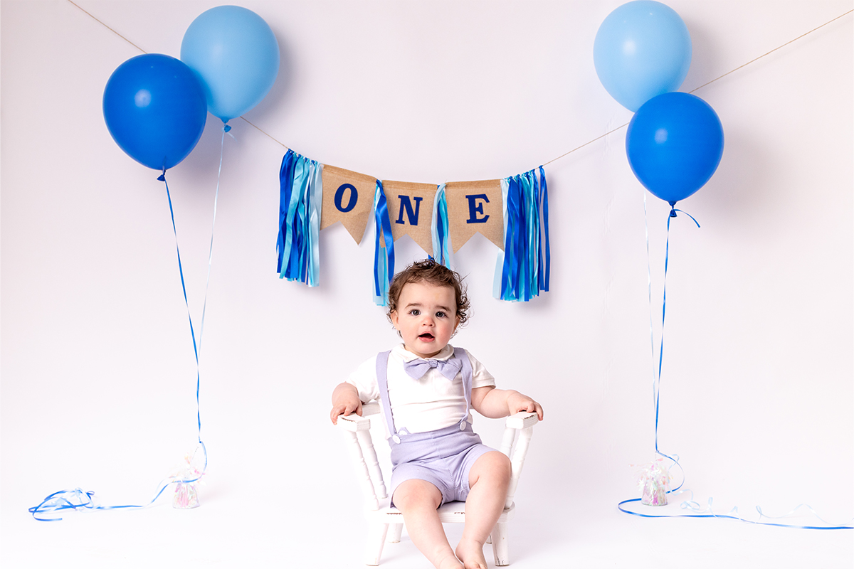 A baby sits on a white chair in front of blue balloons and a "ONE" banner, celebrating a first birthday—captured beautifully by a Long Island photographer.