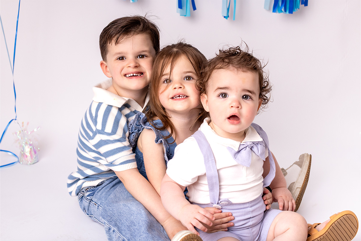 Three young children sit together on a white background, with blue and white decorations behind them. Captured by a Long Island photographer, the older two lovingly hug the baby in front.