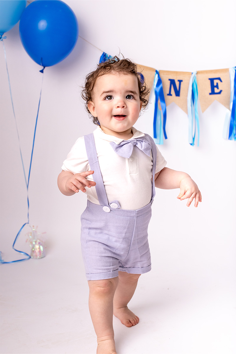 A toddler in a light-colored outfit stands in front of a "ONE" banner with blue balloons in the background, captured perfectly by a Long Island photographer.