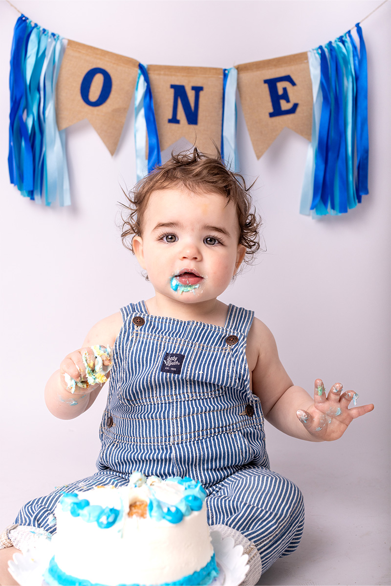 A baby in striped overalls sits in front of a cake with blue and white icing, as blue "ONE" birthday decorations hang in the background—perfectly captured by a Long Island photographer.