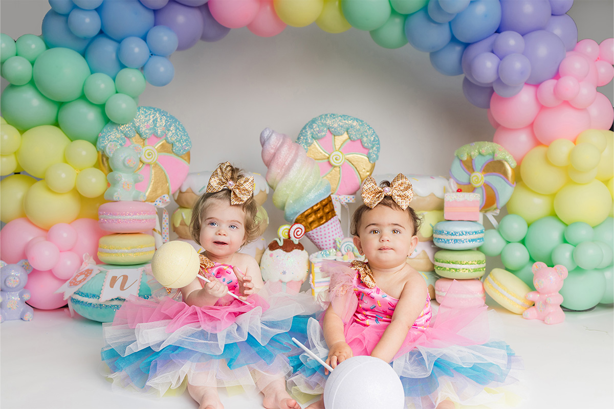 Two babies in colorful tutus and gold bows sit in front of a pastel candy-themed backdrop with balloon decorations, captured perfectly by a Long Island photographer.