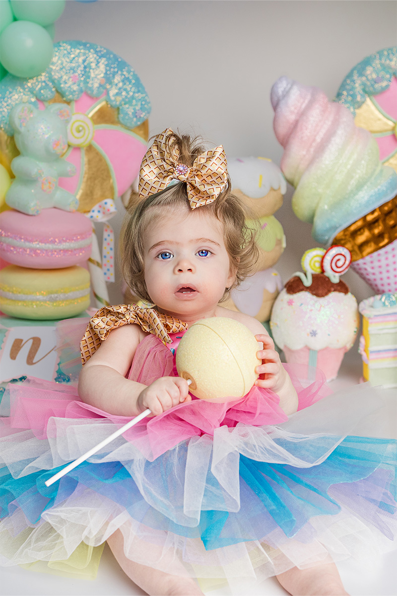 A young child in a colorful tutu and large bow sits surrounded by pastel dessert decorations, holding a large yellow lollipop—captured perfectly by a talented Long Island photographer.
