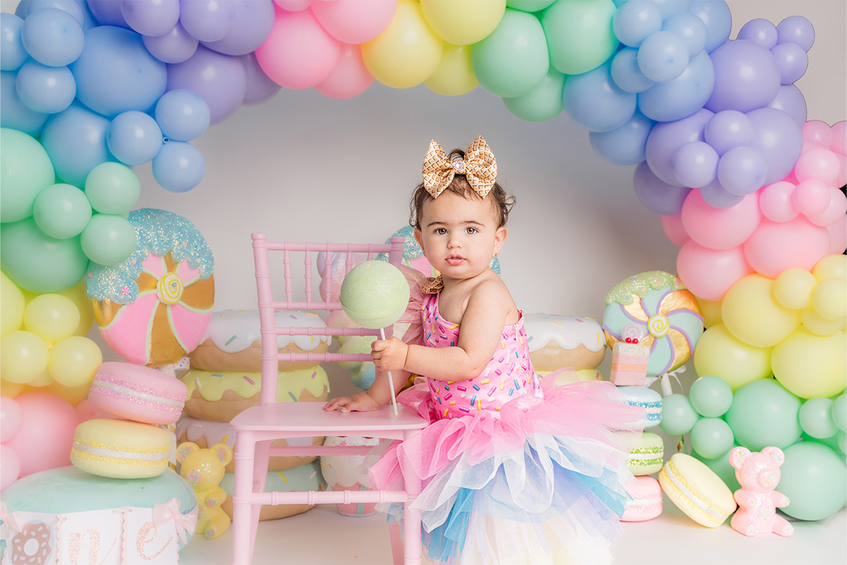 A toddler in a colorful tutu and gold bow headband stands by a pink chair, holding a green lollipop, captured beautifully by a Long Island photographer with pastel balloon arch and candy-themed decorations in the background.