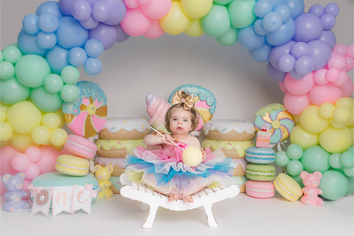 A toddler in a colorful tutu and gold bow sits on a white bench, surrounded by pastel balloons and oversized dessert decorations at a birthday photoshoot captured by a Long Island photographer.
