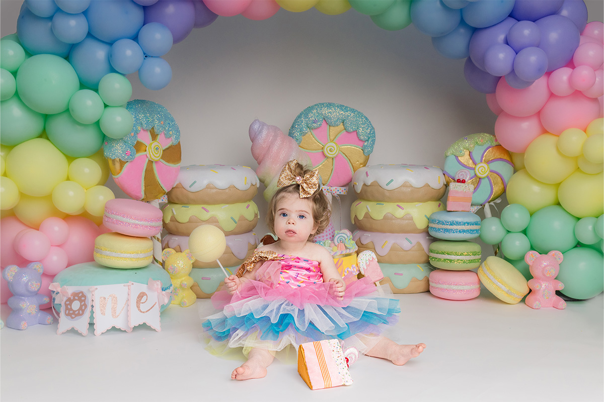 A young child in a colorful tutu sits on the floor holding a balloon, surrounded by pastel balloons and large dessert props in a festive setting, captured perfectly by a Long Island photographer.