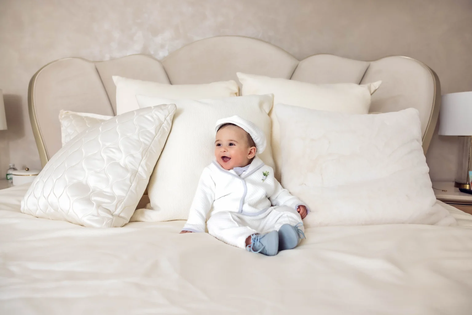 A baby wearing a white outfit and hat sits on a large bed with several white pillows, smiling and looking to the side.