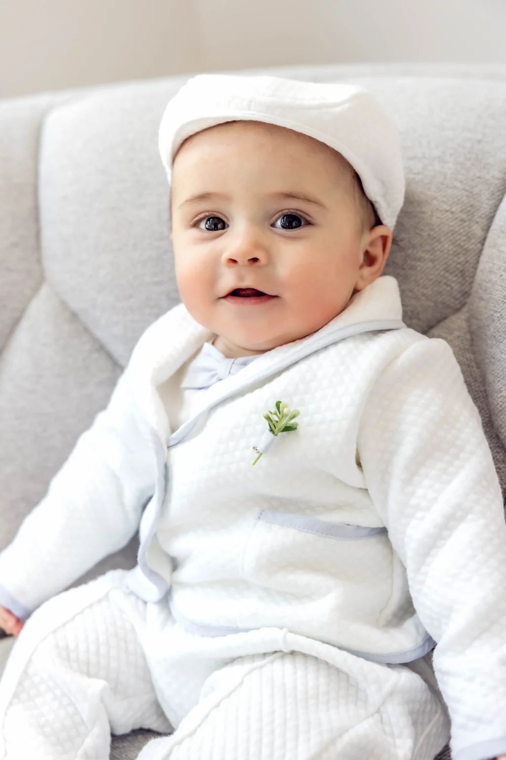 A baby wearing a white textured outfit and a white hat sits on a light gray cushioned chair, looking slightly upward with a neutral expression.