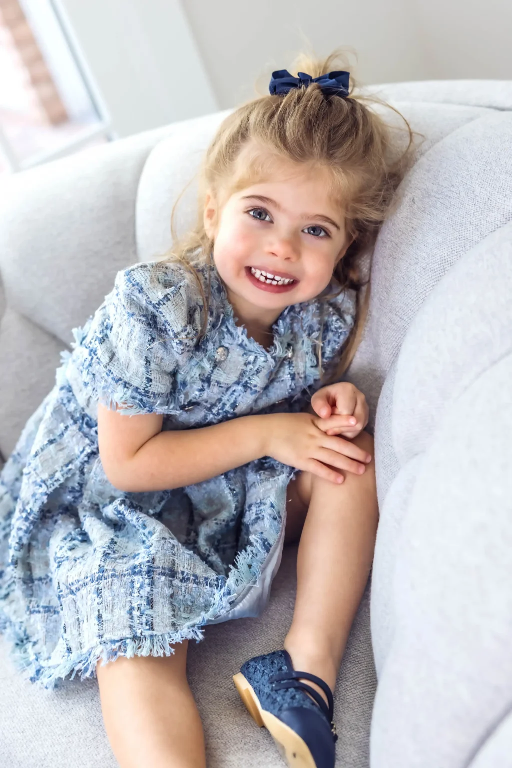 Young girl with a blue bow and patterned dress sits on a light-colored couch, smiling at the camera with hands resting on her knee.
