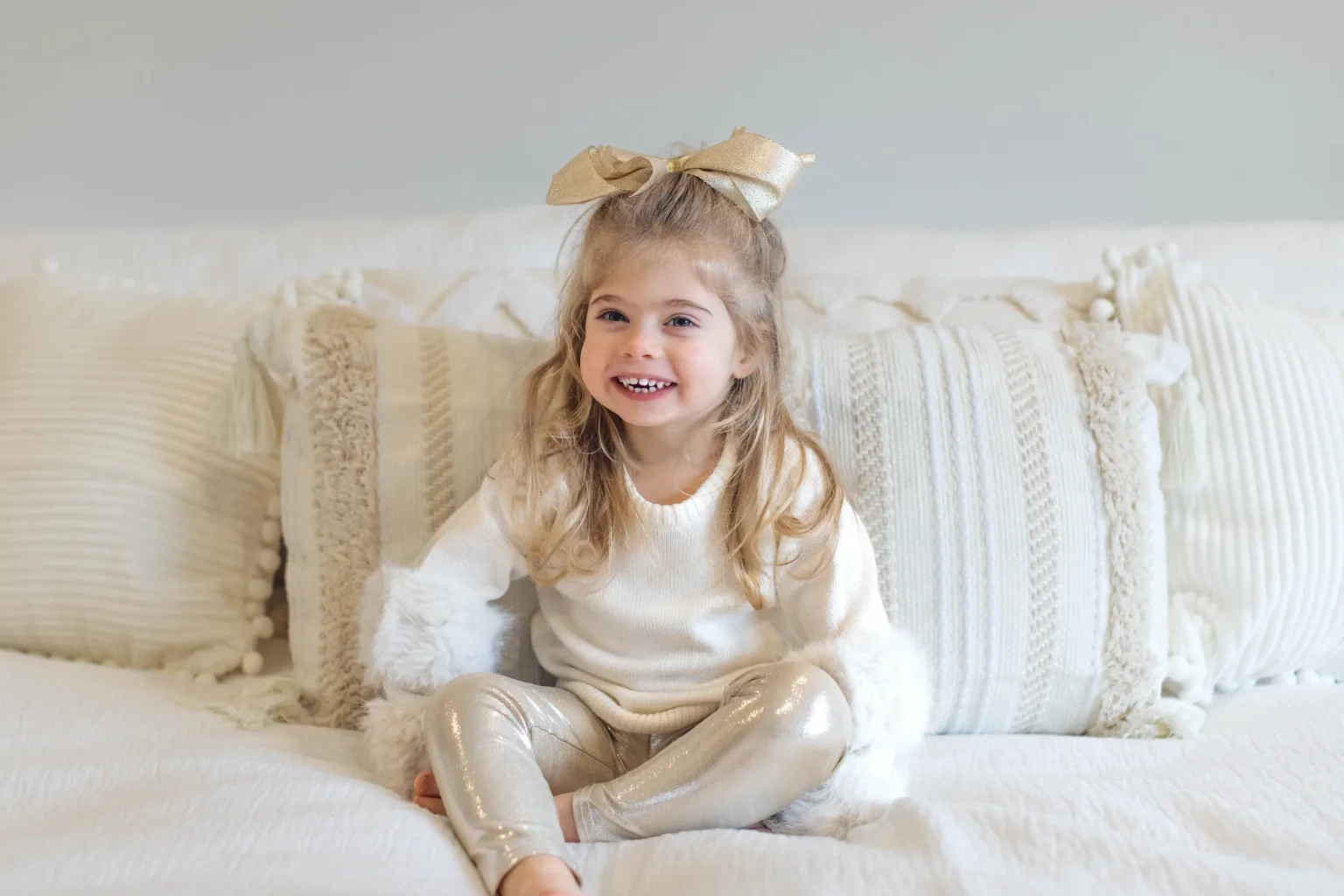 Young girl with long blonde hair, wearing a large gold bow, white sweater, and shiny gold leggings, sits smiling on a white bed with textured pillows.