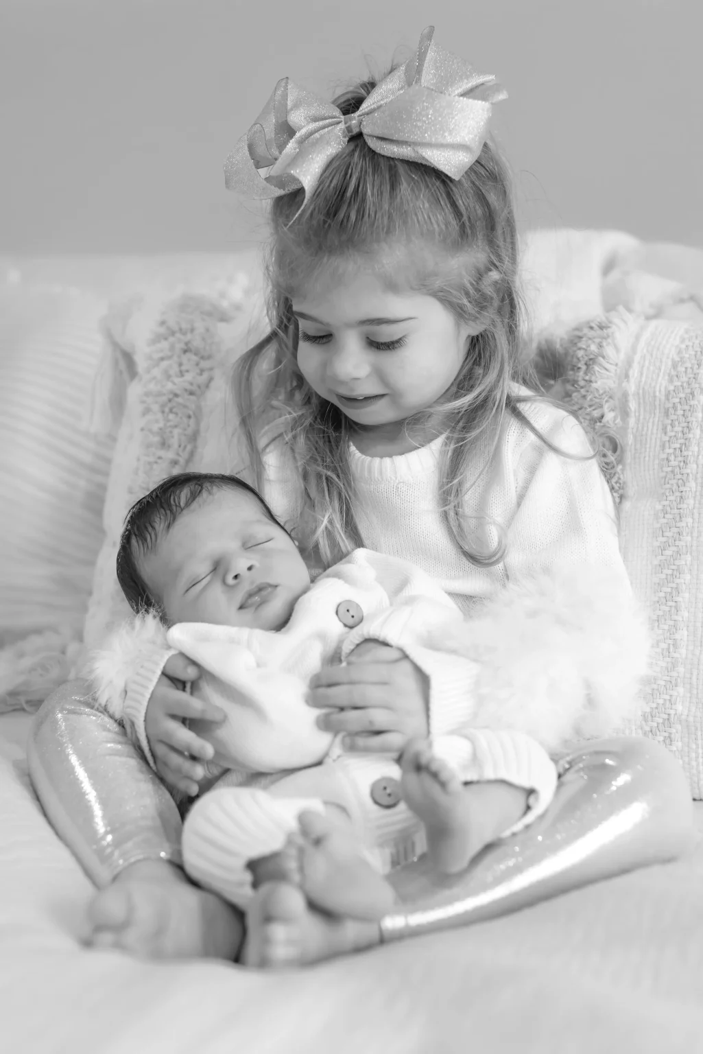 A young girl with a bow in her hair sits on a bed holding a newborn baby in her lap. Both children wear light-colored clothes.