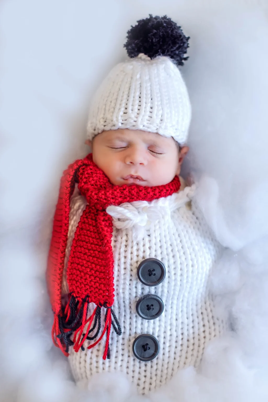 A baby sleeps dressed in a knit snowman outfit with a white hat, red scarf, and black buttons, surrounded by white fluffy material.