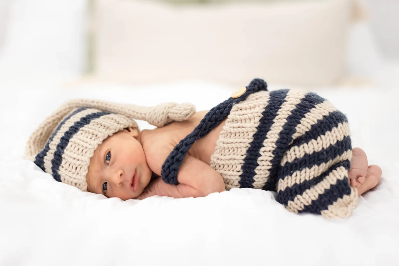 Newborn baby lying on a white bed, dressed in a knitted striped hat and pants in beige and dark blue colors.
