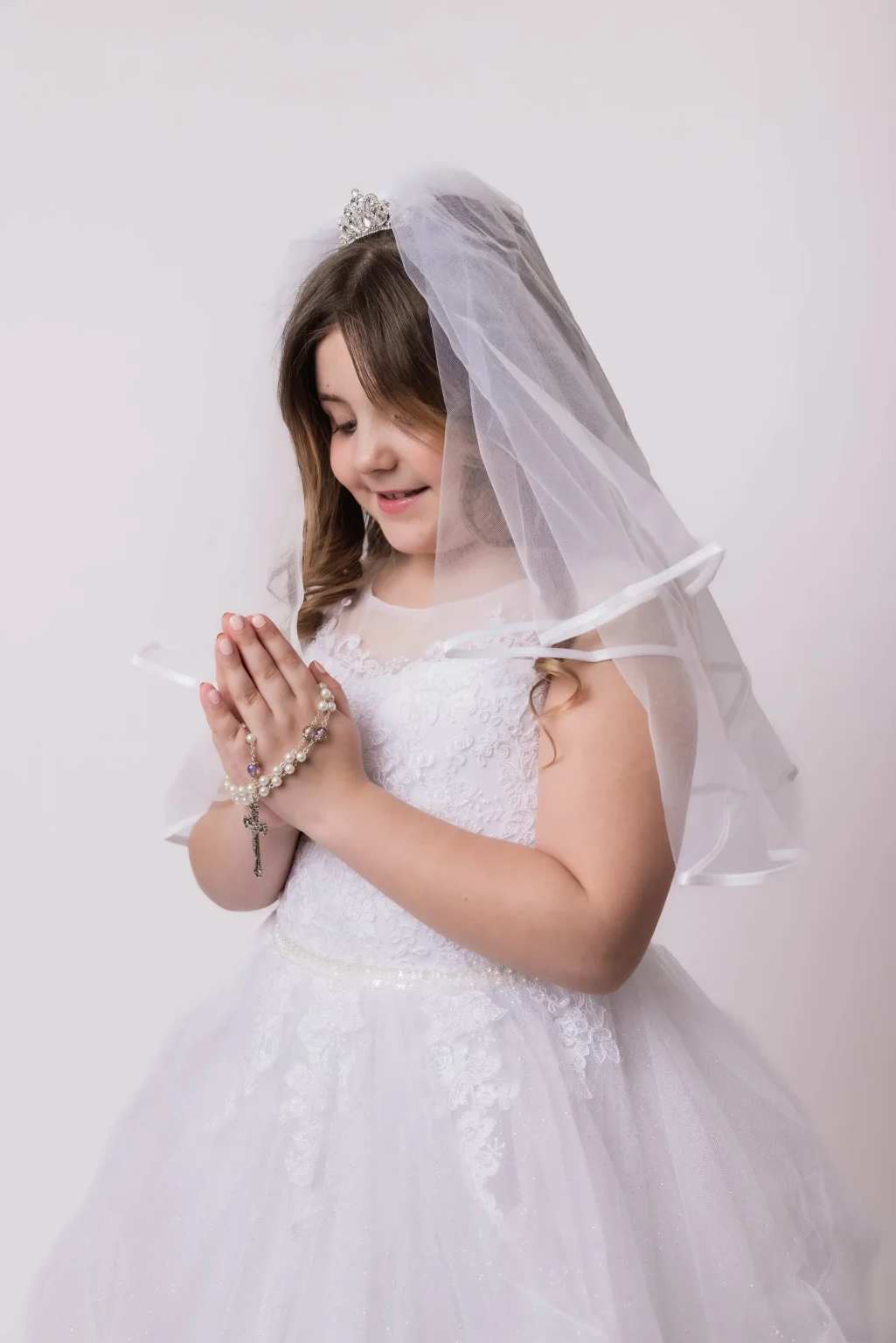 Young girl wearing a white dress and veil, holding a rosary with her hands together in prayer, standing against a plain background.