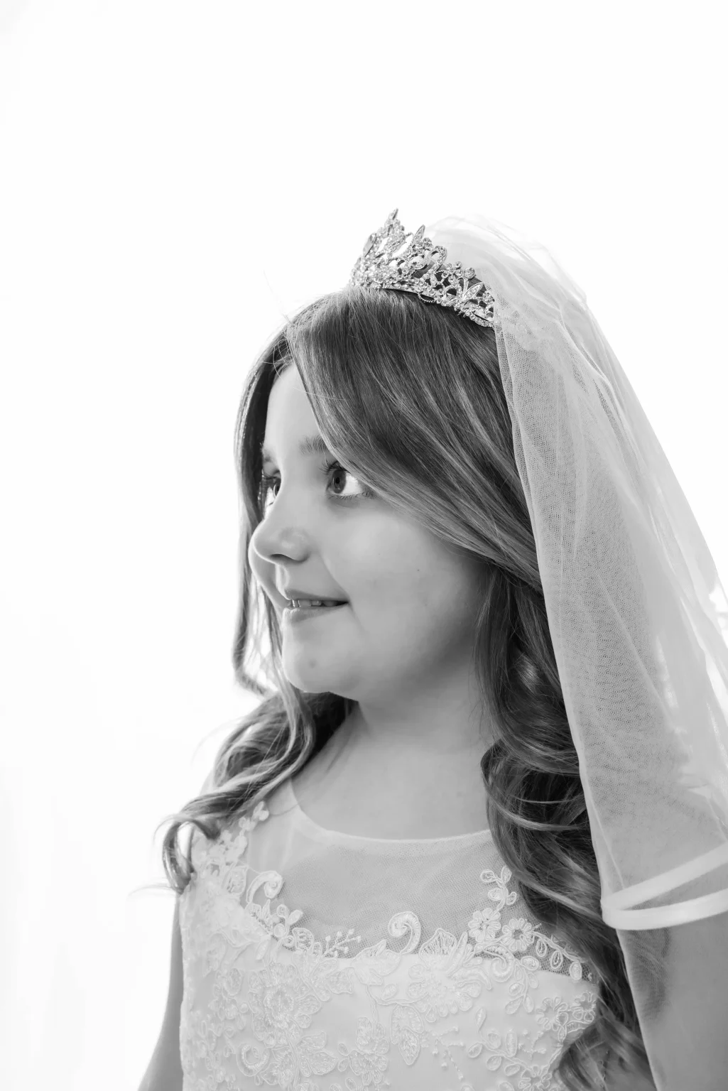 Black and white photo of a young girl wearing a tiara and veil, dressed in a lace-trimmed gown, looking to the side and smiling.