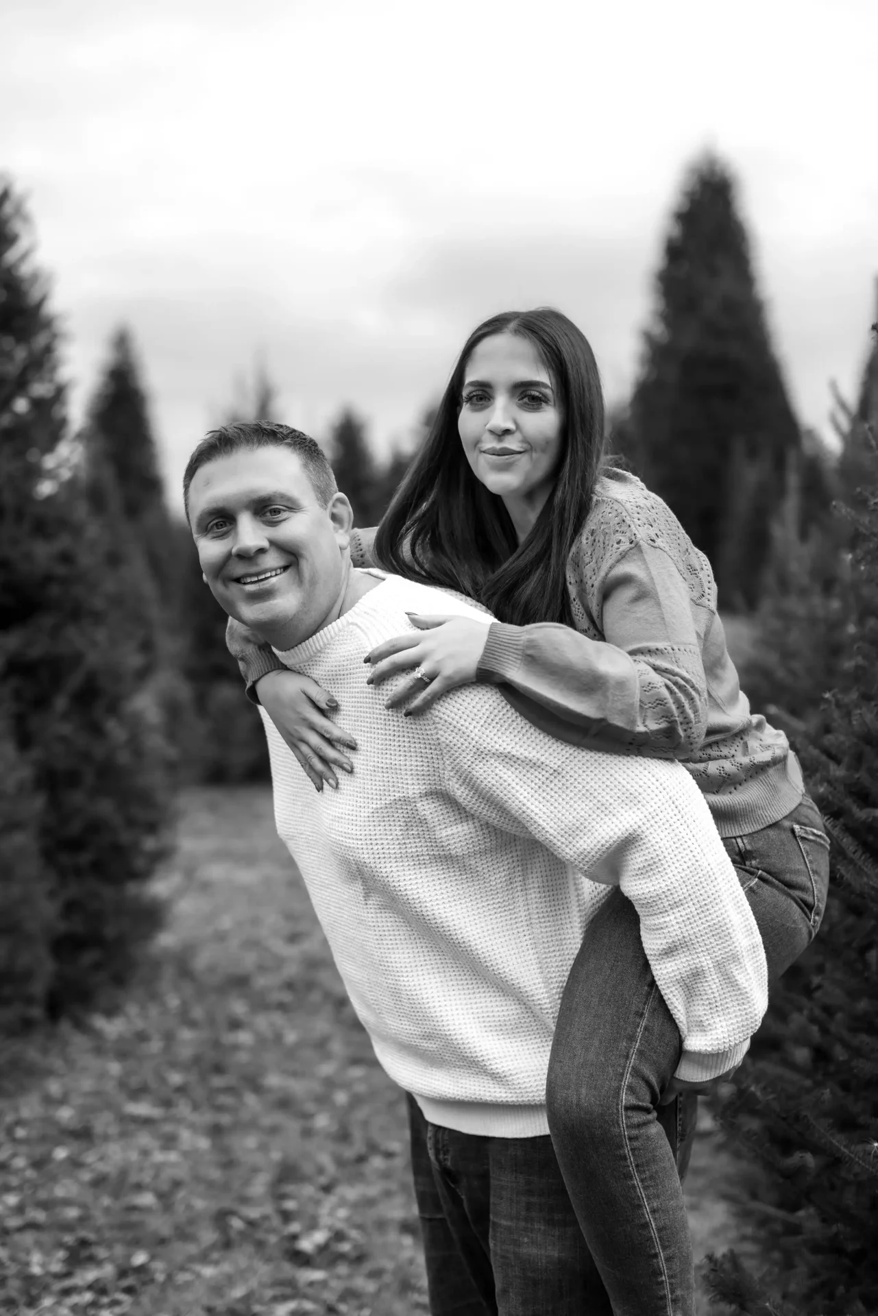 A man gives a woman a piggyback ride outdoors among fir trees, both looking at the camera and smiling. The image is in black and white.