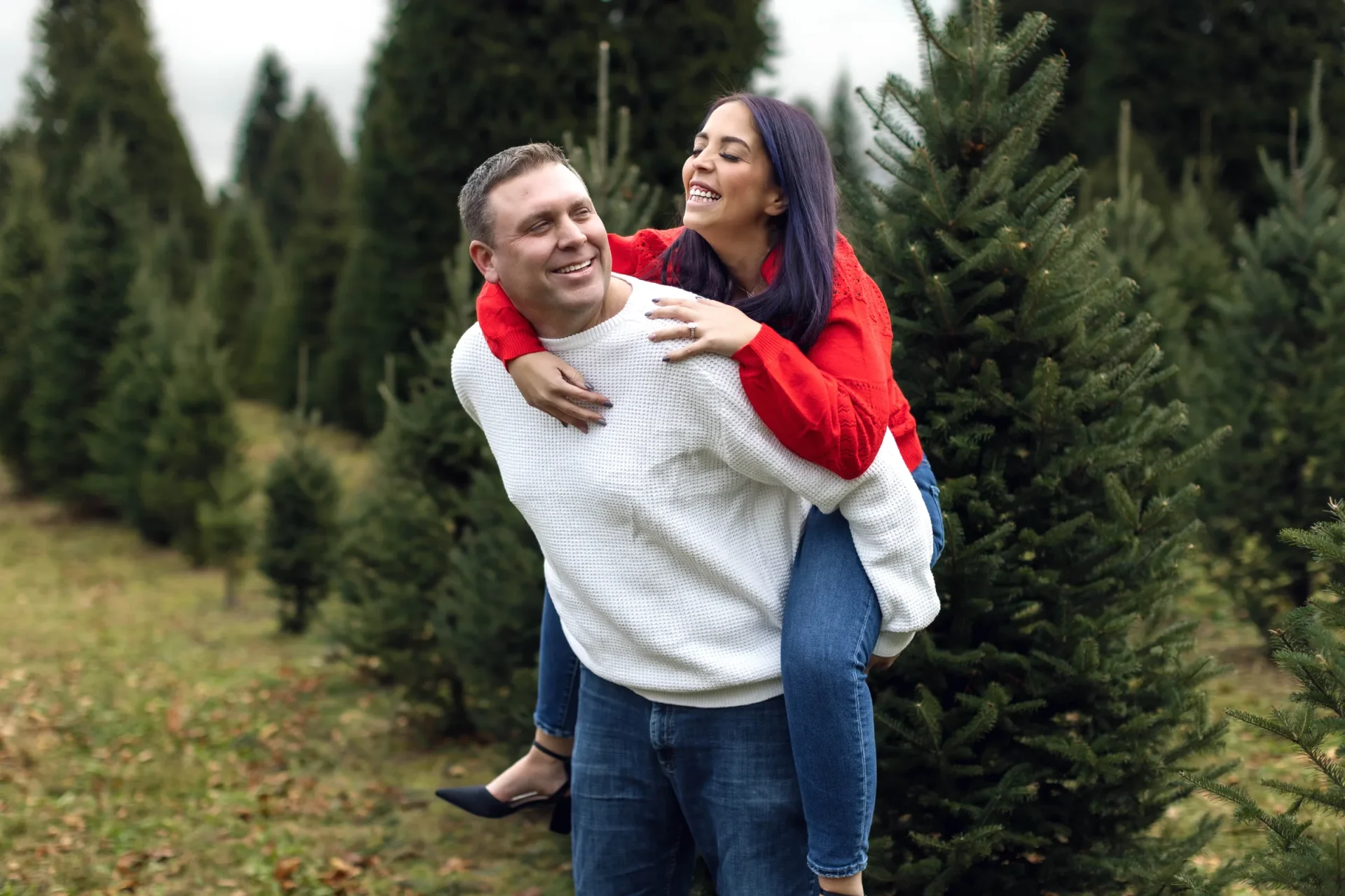 A man giving a woman a piggyback ride outdoors in a field of evergreen trees. Both are smiling and dressed in casual clothes.